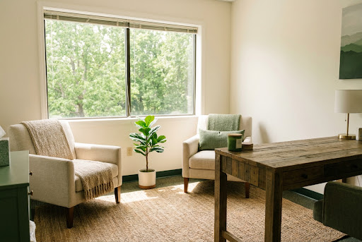 A warm, inviting therapy office featuring a comfortable beige armchair with a textured throw blanket, a rustic wooden desk, and a potted fiddle leaf fig plant near a large window looking out onto lush green trees. The natural light creates a grounded, peaceful refuge.